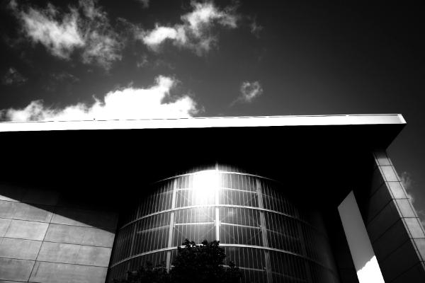 Modernes Gebäude mit zylindrischer Glasfassade unter bewölktem Himmel, fotografiert von unten gegen die Sonne.